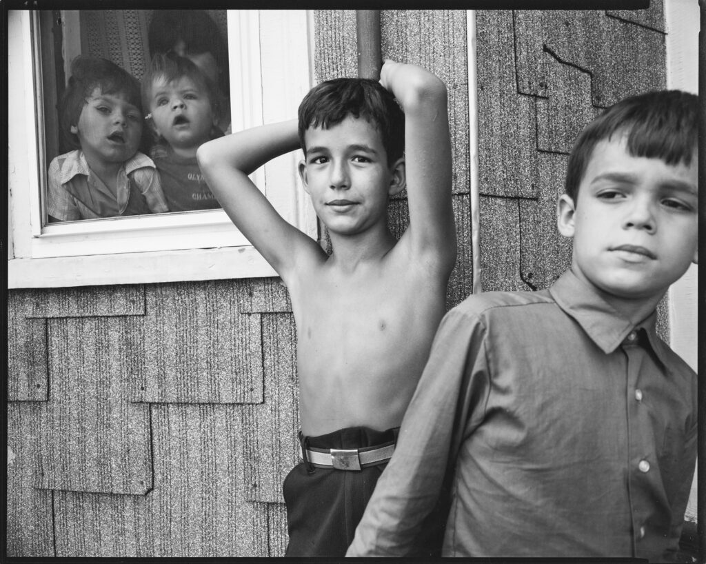 Photographie de N.Nixon de deux jeunes garçons au premier plan et deux enfants plus petits au second plan derriere une fenêtre.