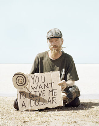 Man with cap sitting on the ground holding a sign that says you want to give me a dollar