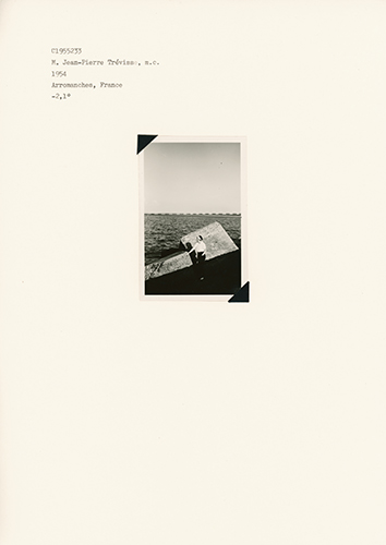 Small B/W photo showing a person in front of stone blocks looking out to sea to the left and with horizon line behind him.